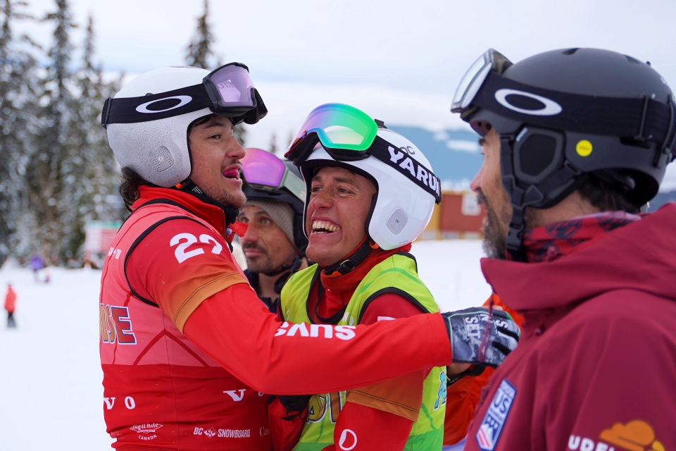 Vier männliche Skifahrer mit Helmen und Skibrillen feiern gemeinsam auf einer verschneiten Piste, zwei von ihnen umarmen sich und lächeln, im Hintergrund sind Bäume und ein Gebäude zu sehen.