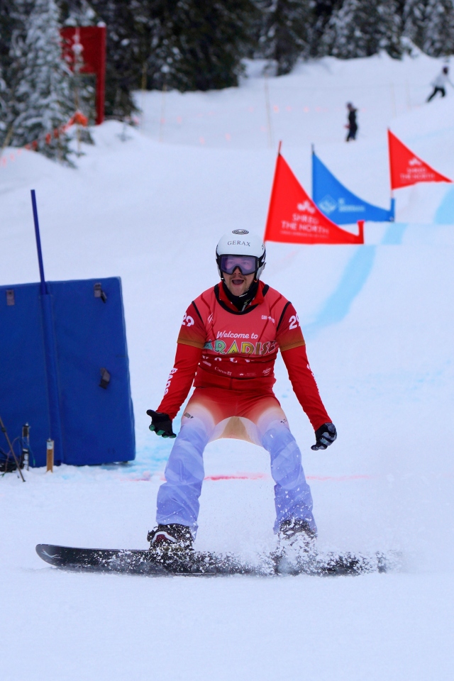 Ein Snowboarder mit roter Jacke, weißem Helm und Skibrille fährt während eines Rennens einen verschneiten Hang hinunter, mit blauen Linien und roten Fahnen im Hintergrund.