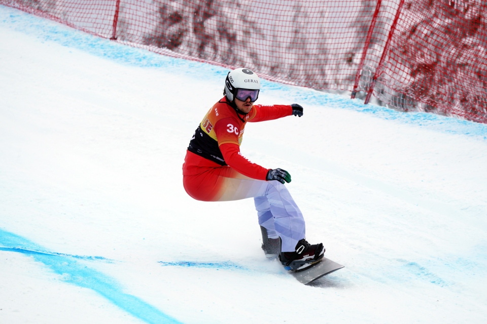 Ein Snowboarder in rot-weißer Ausrüstung macht eine scharfe Kurve auf einer verschneiten Piste, er nimmt an einem Rennen mit nummerierter Startnummer und Helm teil, mit einem roten Sicherheitsnetz im Hintergrund.