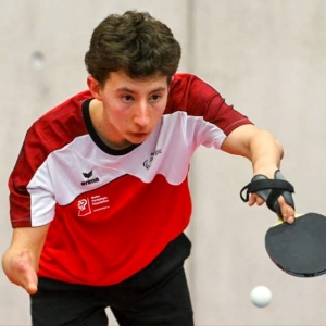 Ein junger Parasportler in einem rot-weißen Hemd spielt intensiv Tischtennis und konzentriert sich darauf, den Ball mit einem schwarzen Schläger zu schlagen, den er in der rechten Hand hält, die mit einem Halteband ausgestattet ist.