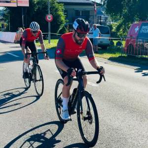 Zwei Radfahrer in roten Trikots und Helmen fahren während eines Rennens auf einer sonnenbeschienenen Straße, während im Hintergrund Veranstaltungsbanner, ein Begleitfahrzeug und Zuschauer zu sehen sind.