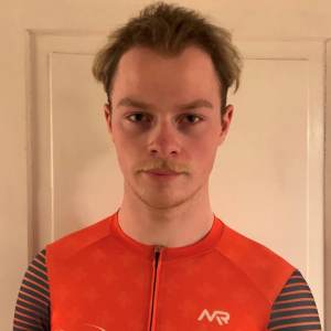 Ein junger Mann mit kurzen, hellbraunen Haaren, der ein orangefarbenes sportliches Oberteil mit NVR-Logo trägt, steht in einem Innenraum vor einer schlichten, hellen Tür.