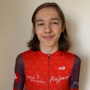 Eine junge Person mit glattem, schulterlangem braunem Haar trägt ein rot-schwarzes Radtrikot mit Logos und steht vor einer schlichten hellen Wand und lächelt leicht in die Kamera.