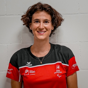 Eine Frau mit kurzen braunen Haaren lächelt in die Kamera. Sie trägt ein rot-schwarz-weißes Trikot der Schweizer Paralympics-Mannschaft und steht vor einer weißen Backsteinwand.