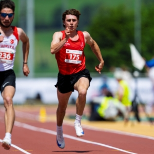 Zwei männliche Athleten sprinten während eines Rennens auf einer Laufbahn. Beide tragen rot-weiße Trikots mit Startnummern; der vordere Läufer trägt die Nummer 175. Im Hintergrund sind Zuschauer und Grünflächen unscharf zu sehen.