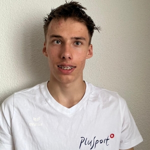 Ein junger Mann mit kurzen, stacheligen braunen Haaren und Zahnspange lächelt in die Kamera. Er trägt ein weißes T-Shirt mit dem PluSport-Logo und steht vor einer schlichten, hellen Wand.
