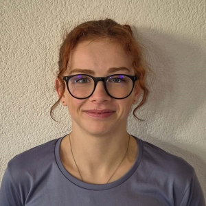 Eine junge Frau mit roten Locken, einer schwarz umrandeten Brille und einem lila Oberteil steht vor einer hellen, strukturierten Wand und lächelt sanft in die Kamera.