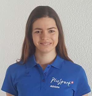 Eine Frau mit langen braunen Haaren und einem blauen Poloshirt mit der Aufschrift „PluSport Athlete“ steht vor einer weißen strukturierten Wand und lächelt in die Kamera.