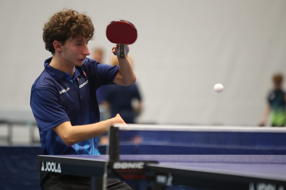 Ein junger männlicher Tischtennisspieler mit lockigem Haar schlägt den Ball auf. Er trägt ein blaues Poloshirt und hält einen roten Schläger. Der Tischtennisball befindet sich in der Luft.
