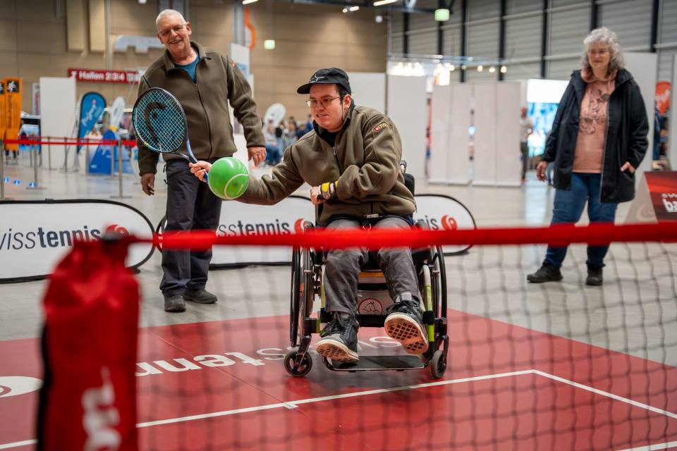 Ein junger Mann im Rollstuhl spielt Tennis mit einem großen grünen Ball und einem Schläger auf einem Hallenplatz, während zwei Erwachsene im Hintergrund lächelnd zusehen. Die Veranstaltung scheint barrierefrei und inklusiv zu sein.