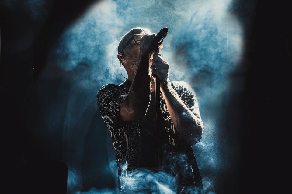 Eine Sängerin mit Mikrofon tritt auf der Bühne auf, umgeben von dramatischem Rauch und stimmungsvoller Beleuchtung. Die Szene ist von hinten beleuchtet, wodurch ein eindrucksvoller Silhouetteneffekt entsteht.