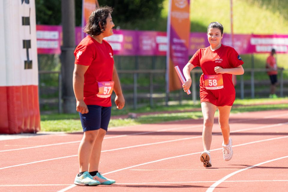 Zwei Sportlerinnen in roten Uniformen befinden sich auf einer Laufbahn. Die eine läuft lächelnd mit einem Staffelstab, während die andere in der Nähe steht und zusieht. Beide tragen Startnummern (57 und 58). Im Hintergrund sind Fahnen und Zäune zu sehen.