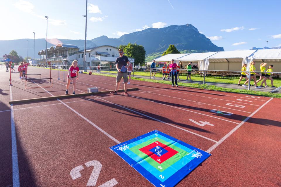 Kinder nehmen an einer sportlichen Aktivität im Freien auf einer Bahn teil und werfen Bälle auf eine bunte Zielmatte. Erwachsene beaufsichtigen sie, im Hintergrund sind Berge, Zelte und andere Menschen an einem sonnigen Tag zu sehen.