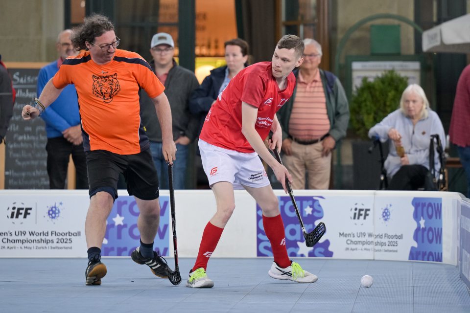 Zwei Männer spielen im Freien Unihockey, einer in einem orangefarbenen und der andere in einem roten Hemd, und kämpfen um den Ball. Die Zuschauer im Hintergrund beobachten das Spiel in der Nähe eines Banners für die Unihockey-Weltmeisterschaft 2025 der Herren U19.
