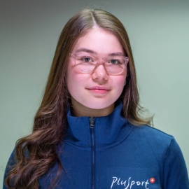 Eine junge Frau mit langen braunen Haaren, die eine Brille und eine blaue Jacke mit dem Plusport-Logo trägt, steht vor einem schlichten hellen Hintergrund, schaut in die Kamera und lächelt leicht.