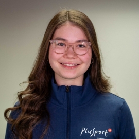 Eine junge Frau mit langen braunen Haaren, die eine Brille und eine blaue Jacke mit dem Plusport-Logo trägt, steht vor einem schlichten hellen Hintergrund, schaut in die Kamera und lächelt leicht.