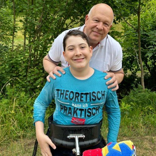 Ein lächelnder Junge im Rollstuhl, der ein blaues Hemd mit deutscher Aufschrift trägt, ist im Freien, hinter ihm steht ein lächelnder älterer Mann, beide umgeben von grünen Bäumen und Laub.