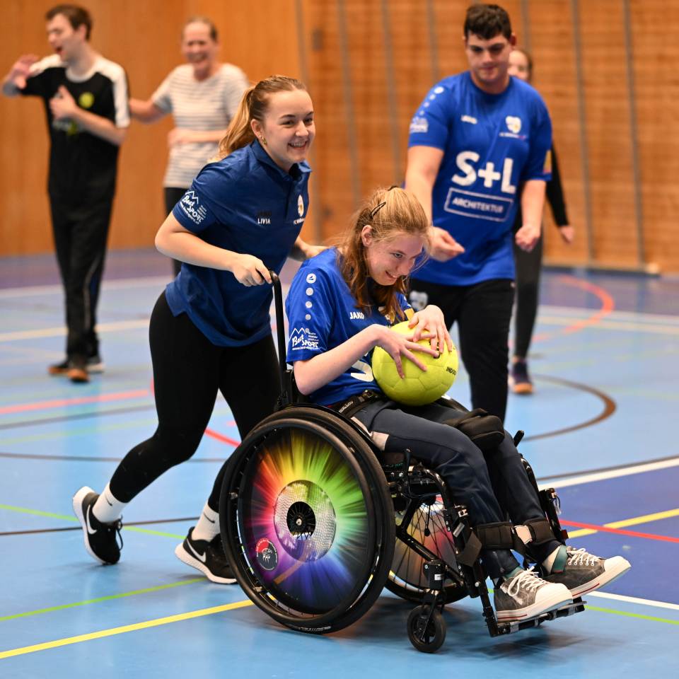 Eine junge Frau in einem bunten Rollstuhl lächelt und hält einen gelben Ball in der Hand. Ihre Teamkollegin im blauen Trikot schiebt ihn auf einem Hallenplatz an. Im Hintergrund jubeln weitere Spielerinnen in blau-weißen Trikots.