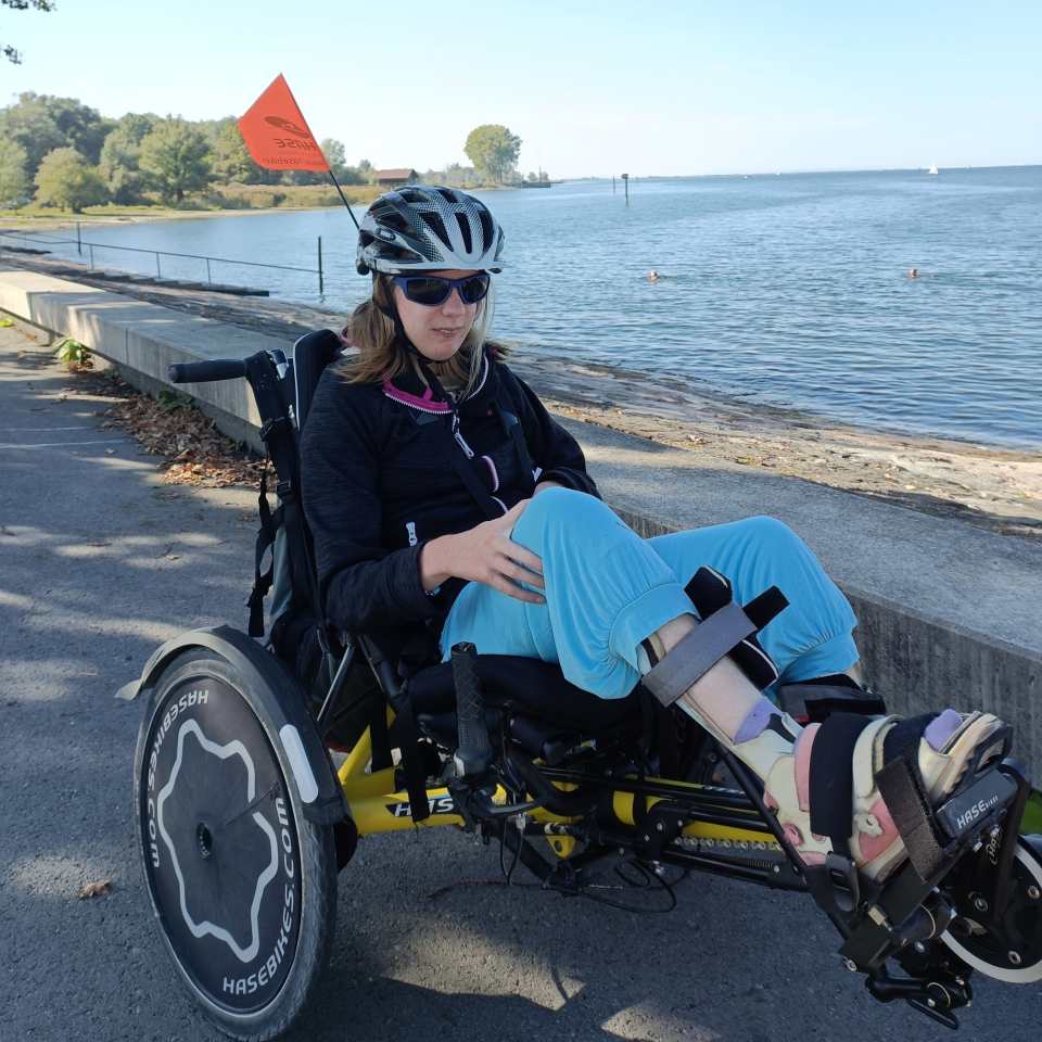 Eine Frau mit Helm, Sonnenbrille und hellblauer Hose fährt mit einem Liegehandbike einen Uferweg entlang. Sie trägt orthopädische Schienen an den Beinen und am Handbike ist eine Sicherheitsfahne befestigt. Im Hintergrund sind Bäume und Wasser zu sehen.