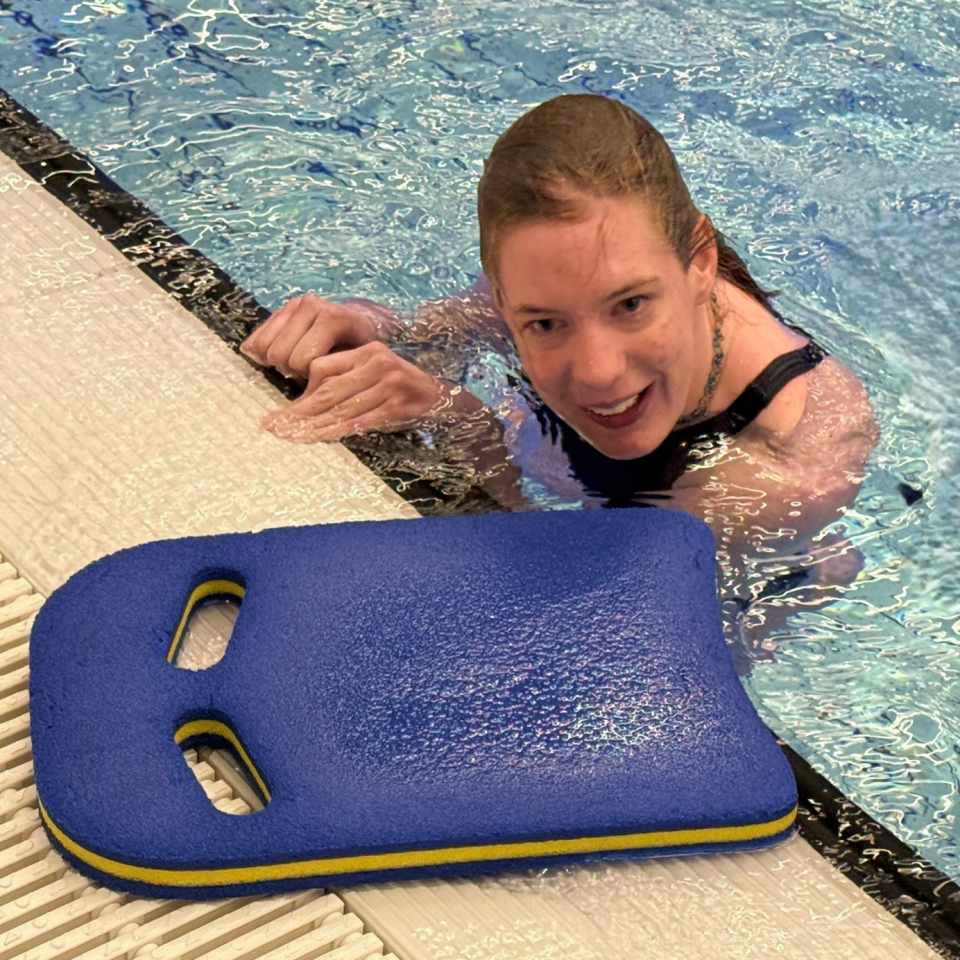 Eine Schwimmerin hält sich am Beckenrand fest und lächelt in die Kamera. Neben ihr liegt ein blaues Schwimmbrett mit zwei Handlöchern am Beckenrand.