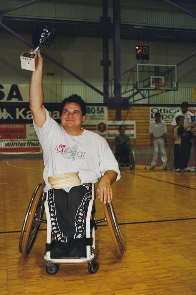 Ein Mann im Rollstuhl reckt einen Pokal in die Höhe und lächelt auf einem Basketballfeld, während er seinen Sieg feiert. Im Hintergrund sind Menschen in der Nähe des Korbs zu sehen.
