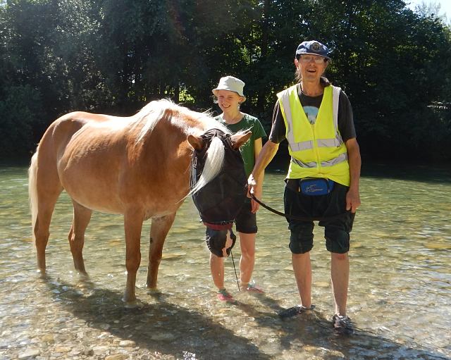 Zwei Personen stehen im flachen Wasser mit einem hellbraunen Pferd. Eine Person trägt eine gelbe Sicherheitsweste und einen Hut, die andere einen Eimerhut. Im Hintergrund sind Bäume und Sonnenlicht zu sehen.