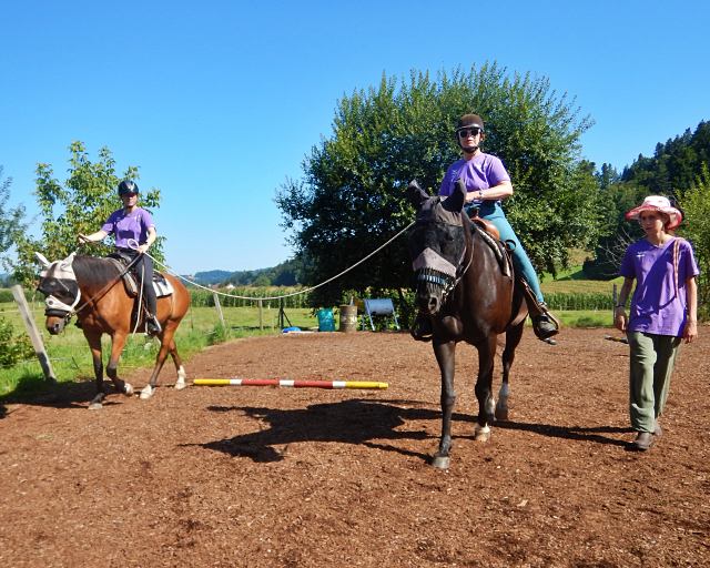 Zwei Personen mit Helmen und lila Hemden reiten auf Pferden in einer Reithalle, während eine weitere Person mit Hut und lila Hemd in der Nähe steht. Im Hintergrund sind Bäume und grüne Hügel unter einem klaren blauen Himmel zu sehen.