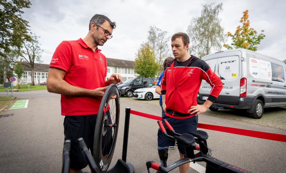 Zwei Männer in roten Hemden stehen neben einem Fahrrad und einem Ersatzrad. Ein Mann erklärt etwas, während der andere zuhört. Im Hintergrund sind ein Lieferwagen und geparkte Autos zu sehen, und ein Mann trägt eine Beinprothese.