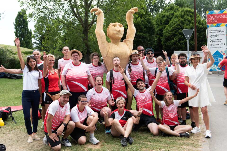 Eine Gruppe von Menschen in passender rot-weißer Sportkleidung posiert lächelnd und jubelnd vor einer großen, hellbraunen Statue einer Figur mit erhobenen Armen im Freien auf einer Wiese mit Bäumen im Hintergrund.