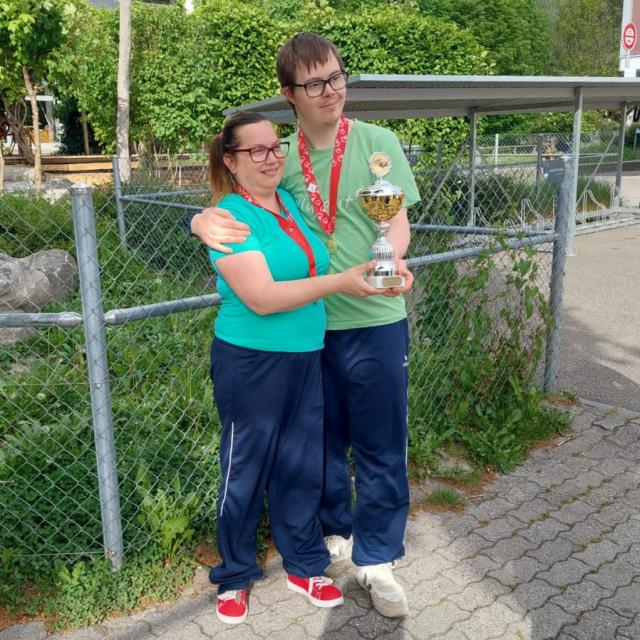 Zwei Personen stehen draußen auf einem gepflasterten Weg, lächeln und halten eine Trophäe in der Hand. Sie tragen beide blaugrüne Hemden, marineblaue Hosen und rote Schuhe. Im Hintergrund sind ein Maschendrahtzaun und grüne Bäume zu sehen.