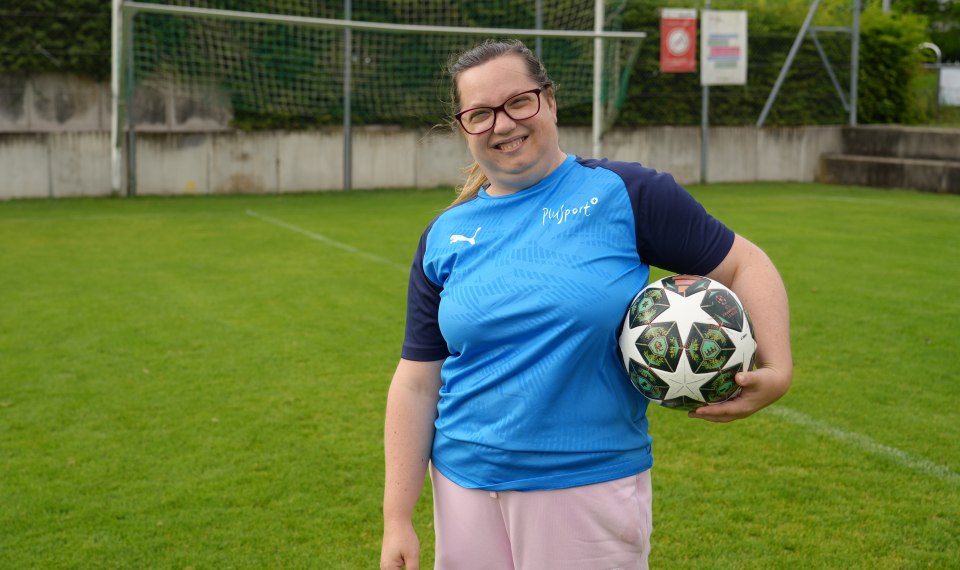Eine Frau mit Brille und blauem Sporthemd steht auf einem Fußballplatz, lächelt und hält einen Fußball unter dem Arm. Im Hintergrund sind der Torpfosten und der grüne Rasen zu sehen.