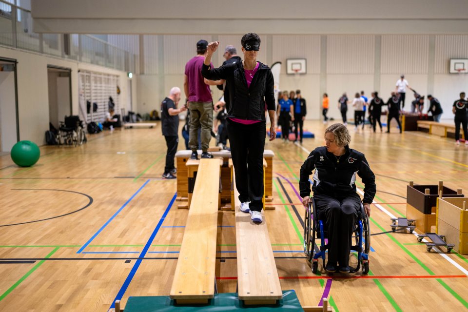 Eine Frau mit einer Augenbinde geht auf einem Schwebebalken, der von einer Frau im Rollstuhl geführt wird, in einer Turnhalle, in der andere Personen an verschiedenen Aktivitäten im Hintergrund teilnehmen.