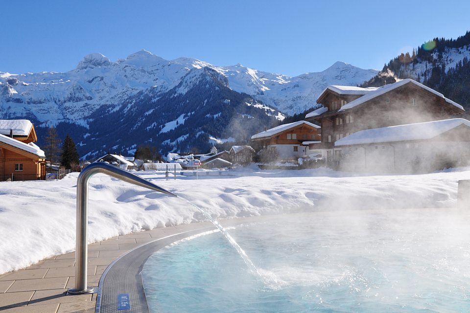 Whirlpool im Freien mit dampfendem Wasser, umgeben von Schnee, Holzhütten und Kiefern, vor der Kulisse verschneiter Berge und unter einem strahlend blauen Himmel.
