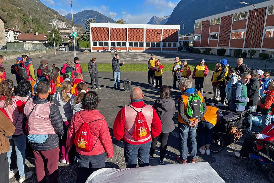 Eine Gruppe von Menschen mit bunten Westen und Rucksäcken versammelt sich in einem sonnigen Außenbereich in der Nähe eines Schulgebäudes und hört einem Redner zu. Im Hintergrund sind Berge und blauer Himmel zu sehen.