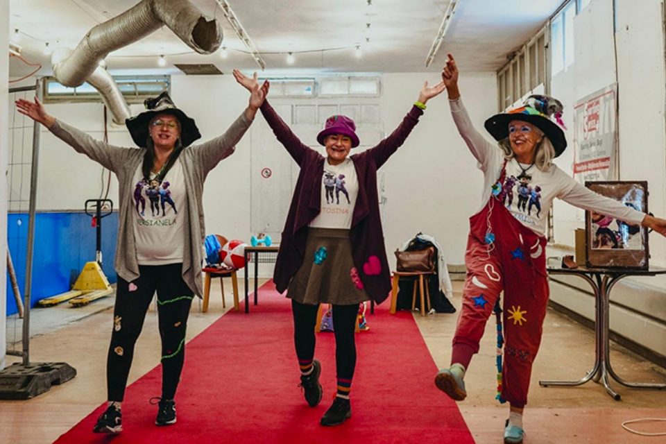 Drei fröhliche Frauen in farbenfrohen, verspielten Outfits und Hüten posieren mit erhobenen Armen und hochgelegten Beinen auf einem roten Teppich in einem hellen Raum, lächeln und feiern gemeinsam.