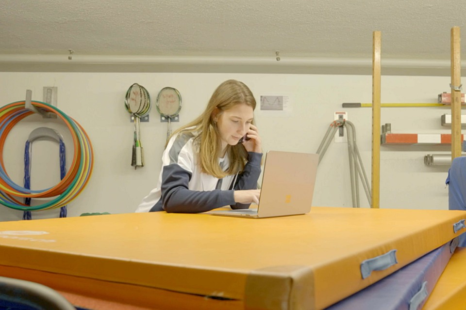 Eine junge Frau sitzt an einem Tisch mit einer orangefarbenen Matte, arbeitet an einem Laptop und telefoniert in einem Raum mit Sportgeräten wie Schlägern und Schläuchen an der Wand hinter ihr.