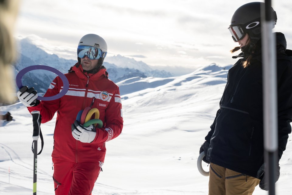Ein Skilehrer in einem roten Anzug mit bunten Ringen unterhält sich mit einem Skifahrer in schwarzer Ausrüstung auf einem verschneiten Berg mit schneebedeckten Gipfeln im Hintergrund unter einem bewölkten Himmel.