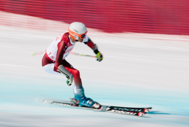 Ein Skifahrer in einem rot-weißen Anzug und einem orangefarbenen Helm rast mit hoher Geschwindigkeit bergab, wobei Bewegungsunschärfe die schnelle Bewegung und ein rotes Sicherheitsnetz im Hintergrund unterstreicht.