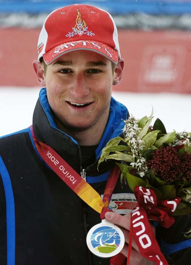 Ein lächelnder Sportler mit roter Mütze und Jacke hält einen Blumenstrauß und zeigt eine Silbermedaille von den Paralympischen Winterspielen 2006 in Turin. Auf seiner Mütze sind Schneeflocken zu sehen, und der Hintergrund ist unscharf.