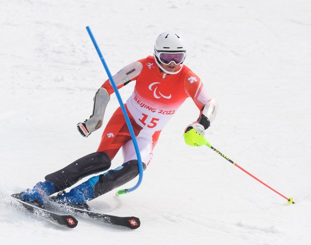 Ein paralympischer Skifahrer in rot-weißer Ausrüstung, mit Brille und Helm, fährt während eines Slalomrennens bei den Paralympischen Winterspielen 2022 in Peking um eine blaue Stange herum bergab.