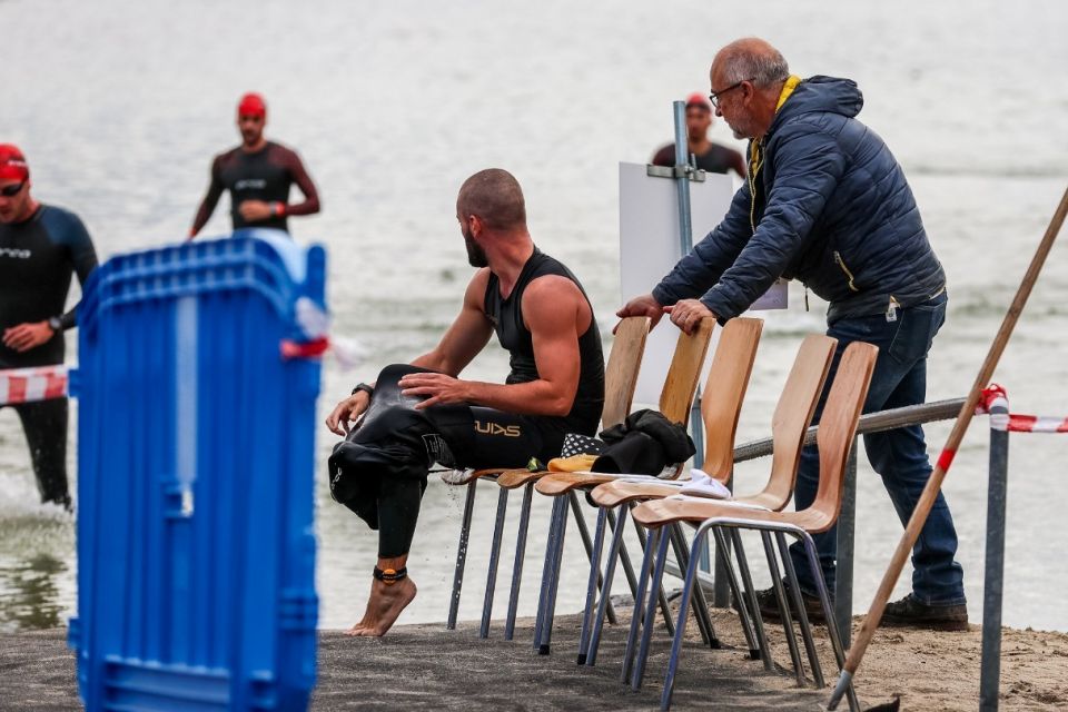 Ein Mann in einem Neoprenanzug sitzt auf einer Reihe von Stühlen am Wasser und zieht seine Ausrüstung aus, während ein anderer Mann in einer Jacke in der Nähe steht. Im Hintergrund tauchen Schwimmer in Neoprenanzügen aus dem Wasser auf. Auf der linken Seite ist eine blaue Plastikbarriere zu sehen.
