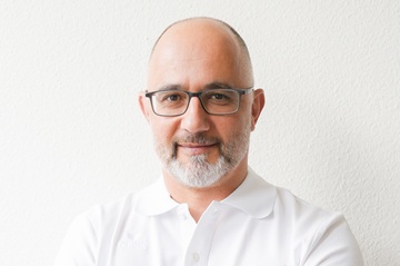 Ein Mann mittleren Alters mit Glatze, grauem Bart und Brille, der ein weißes Hemd mit Kragen trägt, steht vor einer schlichten weißen Wand und lächelt sanft in die Kamera.