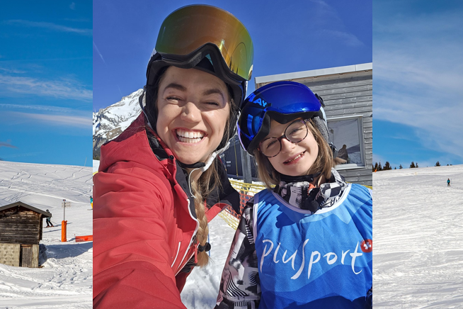 Zwei Personen mit Skihelmen und -brillen lächeln für ein Selfie auf einer verschneiten Skipiste, hinter ihnen eine Holzhütte und ein modernes Gebäude unter einem strahlend blauen Himmel.
