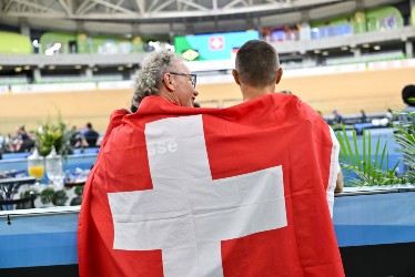 Zwei in eine Schweizer Fahne gehüllte Personen stehen nebeneinander in einem Stadion, von der Kamera abgewandt. Der Veranstaltungsort scheint ein Innenraum mit Sitzgelegenheiten und einer Radrennbahn im Hintergrund zu sein.