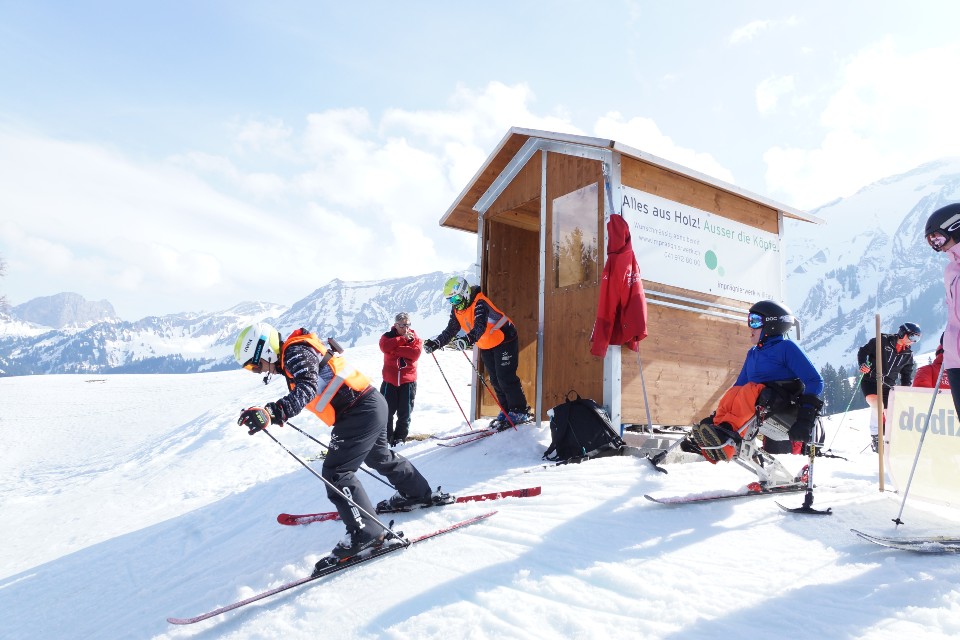 Kinder mit Helmen und orangefarbenen Westen starten ein Skirennen in der Nähe eines kleinen Holzschuppens an einem verschneiten Berghang, mit Lehrern und Bergen im Hintergrund unter einem klaren Himmel.