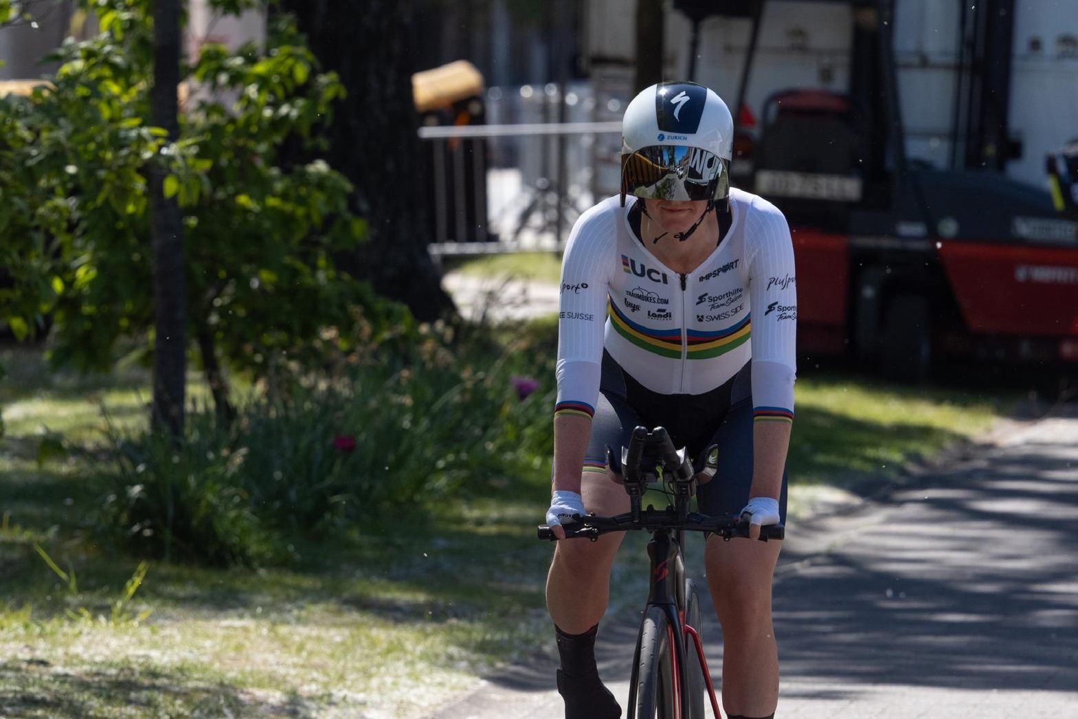 Ein Radfahrer, der ein weißes UCI-Trikot, einen Helm und eine Sonnenbrille trägt, fährt an einem sonnigen Tag mit grünem Laub und einer unscharfen Außenlandschaft im Hintergrund Fahrrad.