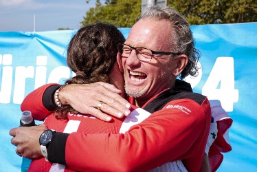 Ein Mann mit Brille und rotem Hemd lächelt freudig, während er eine andere Person, ebenfalls in Rot, bei einer Veranstaltung im Freien umarmt. Im Hintergrund sind ein blaues Schild und Bäume zu sehen.